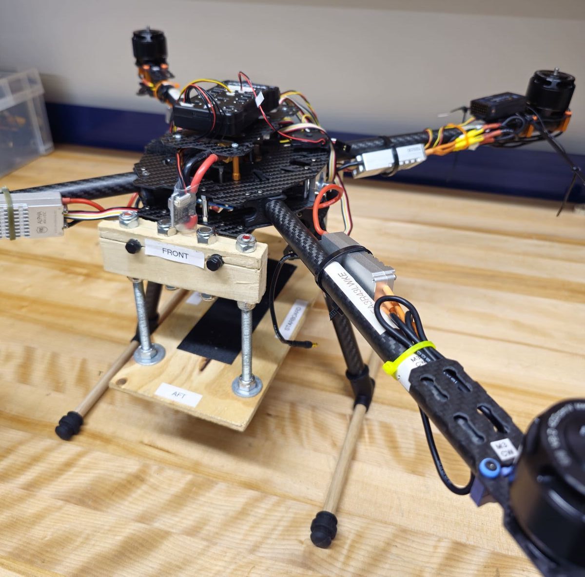 Quadcopter frame on a workbench, with motors, wiring and a wooden mount marked front and aft